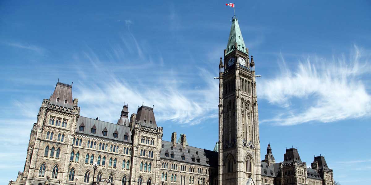 Centre Block of Canada's Parliament Buildings in Ottawa, showcasing the Peace Tower with a green copper roof and Canadian flag at the top. The Gothic Revival architecture features pointed arches, intricate stonework, and steeply pitched roofs. The stone façade includes numerous windows and decorative elements.