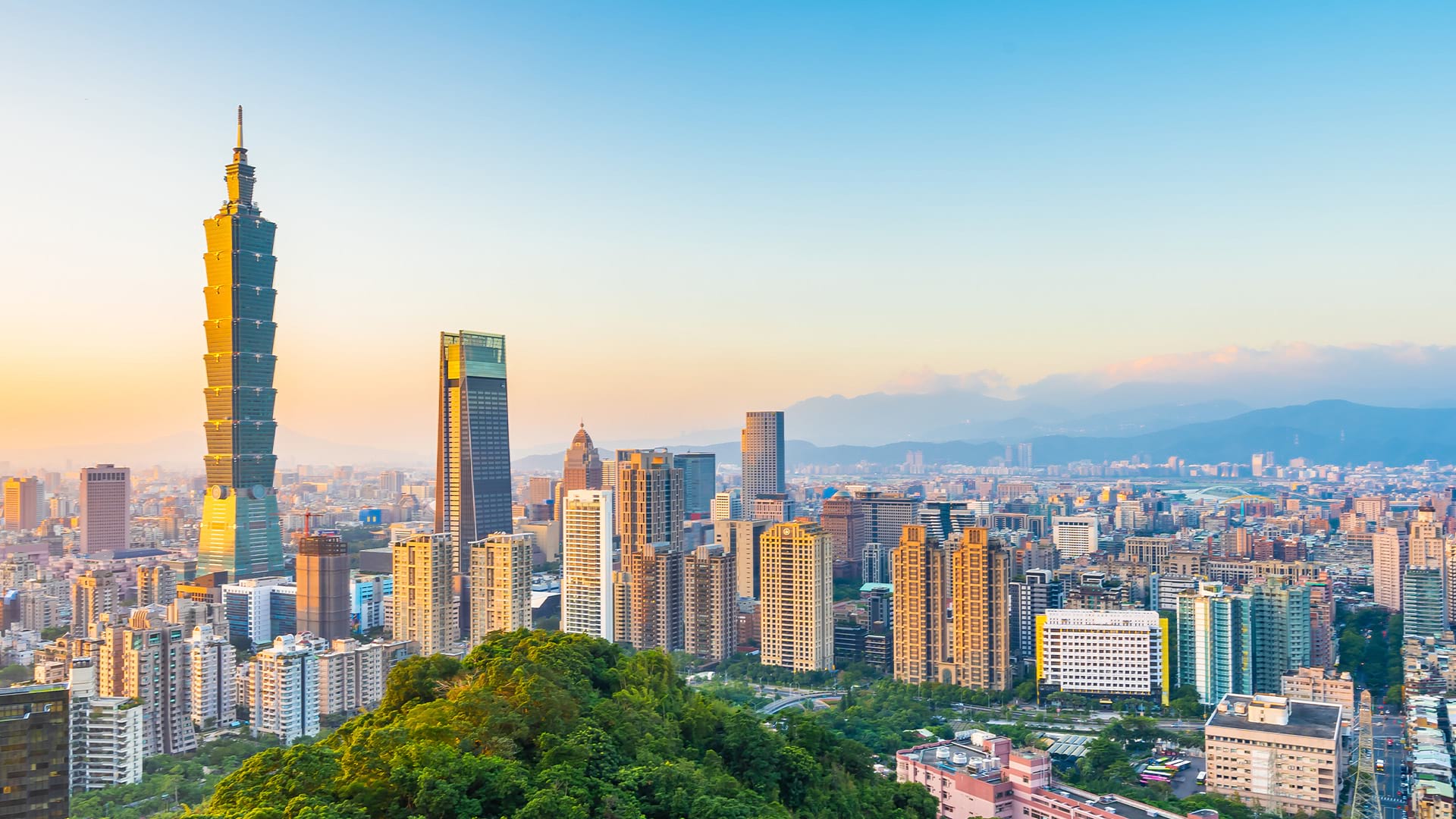 Vue panoramique de la ligne d’horizon de Taipei au coucher du soleil, mettant en vedette le gratte-ciel Taipei 101 entouré de tours modernes, avec des collines verdoyantes au premier plan et des montagnes en arrière-plan sous un ciel dégagé.