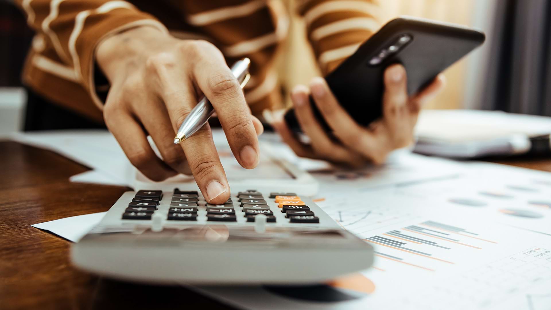 A person is using a calculator while holding a smartphone, surrounded by documents with printed charts and graphs on a desk, suggesting financial planning or budgeting work.