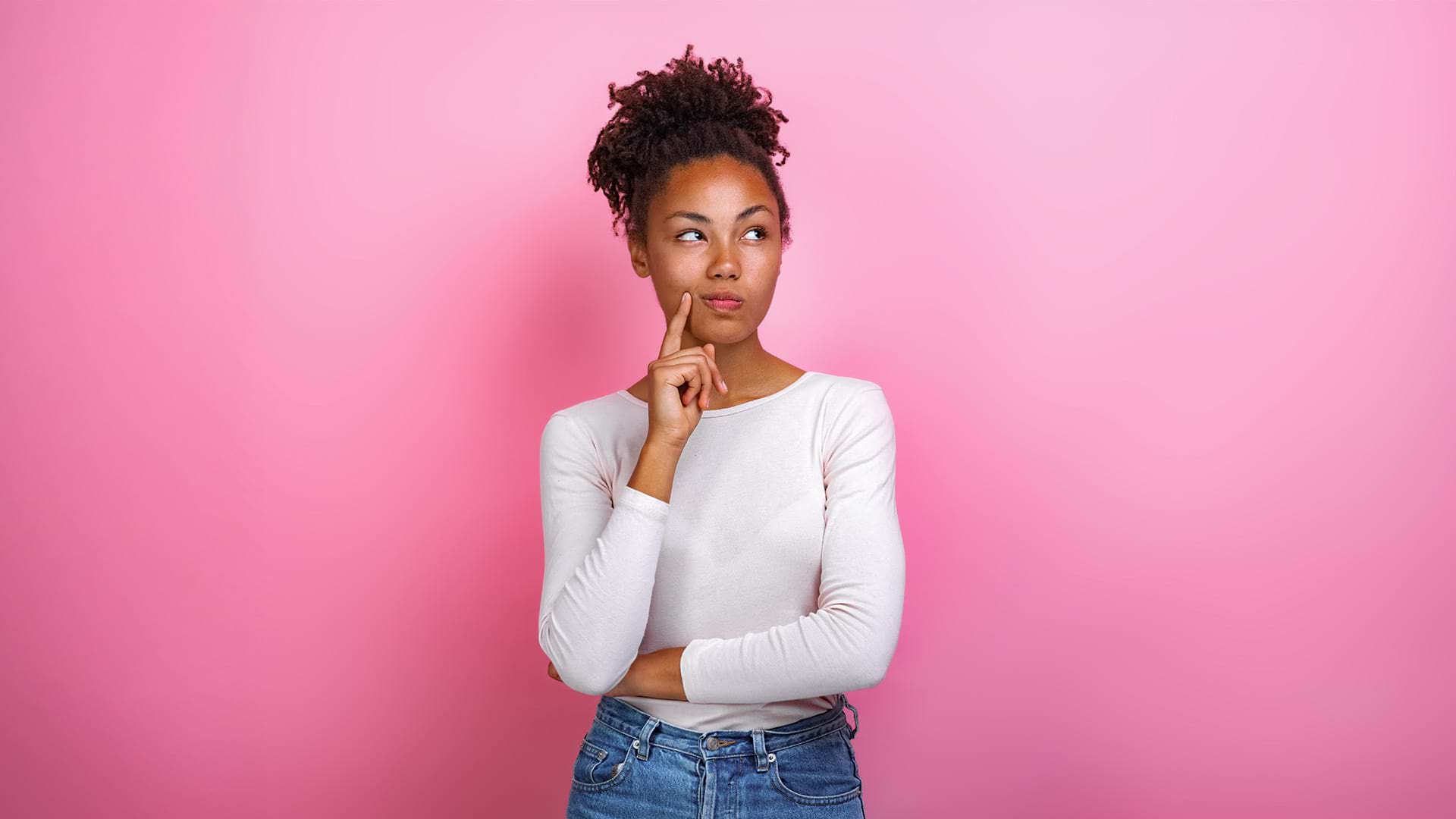 Person standing against a solid pink background, wearing a white long-sleeve shirt and blue jeans. They have curly hair styled in a bun and are posing thoughtfully with one hand on their chin and the other arm crossed.