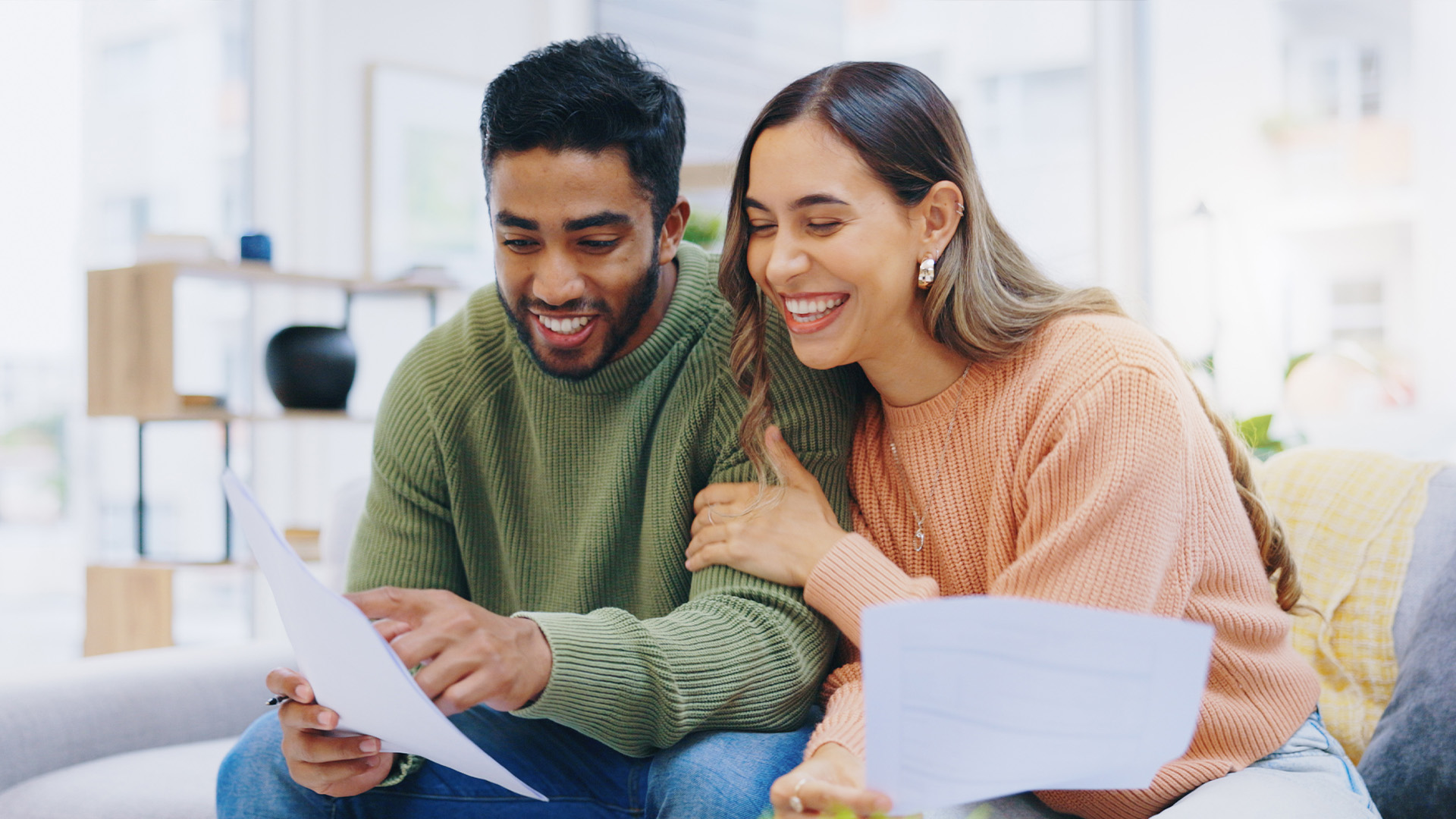 Un couple souriant assis ensemble sur un divan, en train d'examiner des documents. L'homme pointe avec un stylo un papier qu'il tient, tandis que la femme pose affectueusement sa main sur son bras et tient un autre document, tous deux semblant heureux.