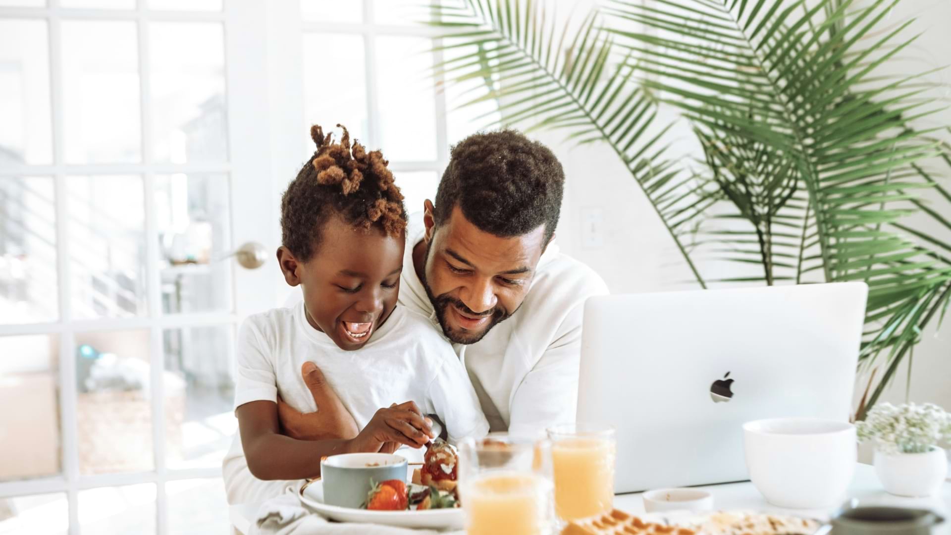 Un enfant et un adulte sont assis ensemble à une table de déjeuner, partageant un repas et regardant une assiette de fruits. Un ordinateur portable, des verres de jus d’orange et une plante d’intérieur sont visibles dans une pièce lumineuse et ensoleillée.