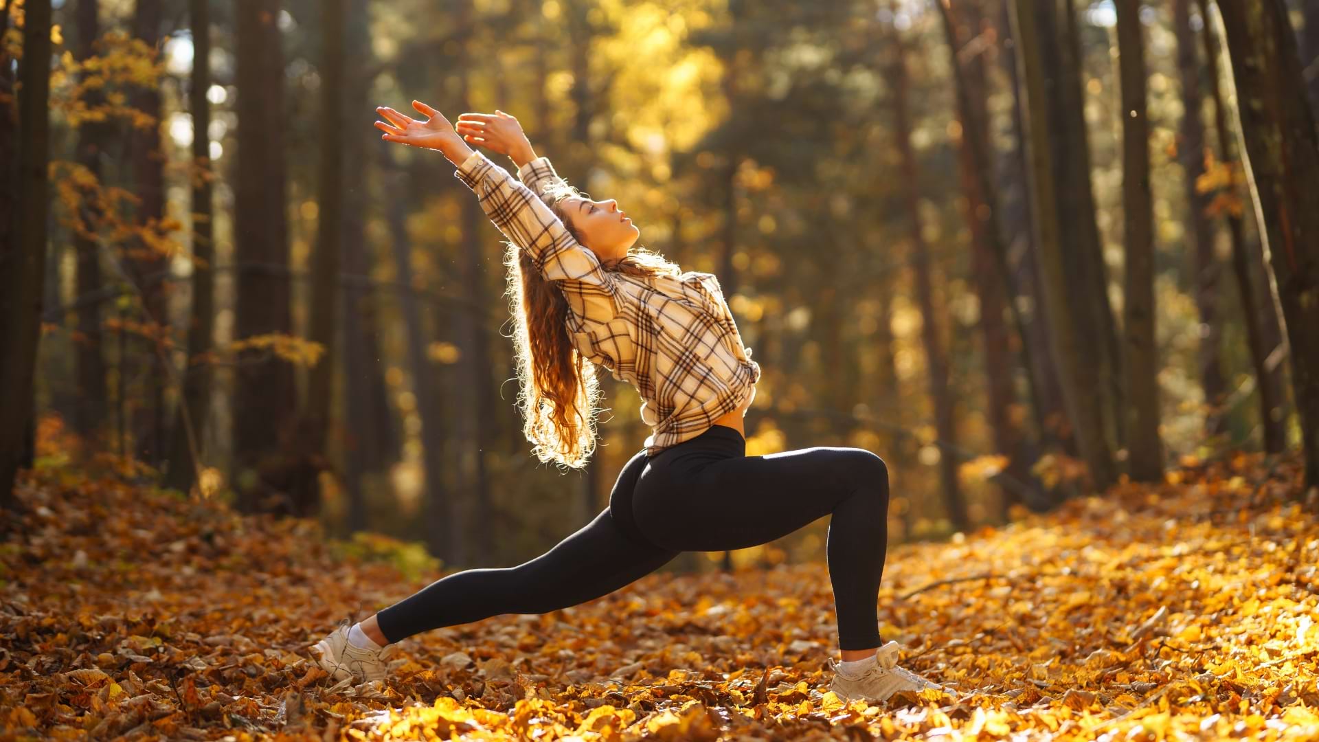 Une personne effectuant une posture de fente de yoga dans une forêt recouverte de feuilles d’automne, les bras étendus au-dessus de la tête. Une lumière dorée traverse les arbres et éclaire le paysage forestier.